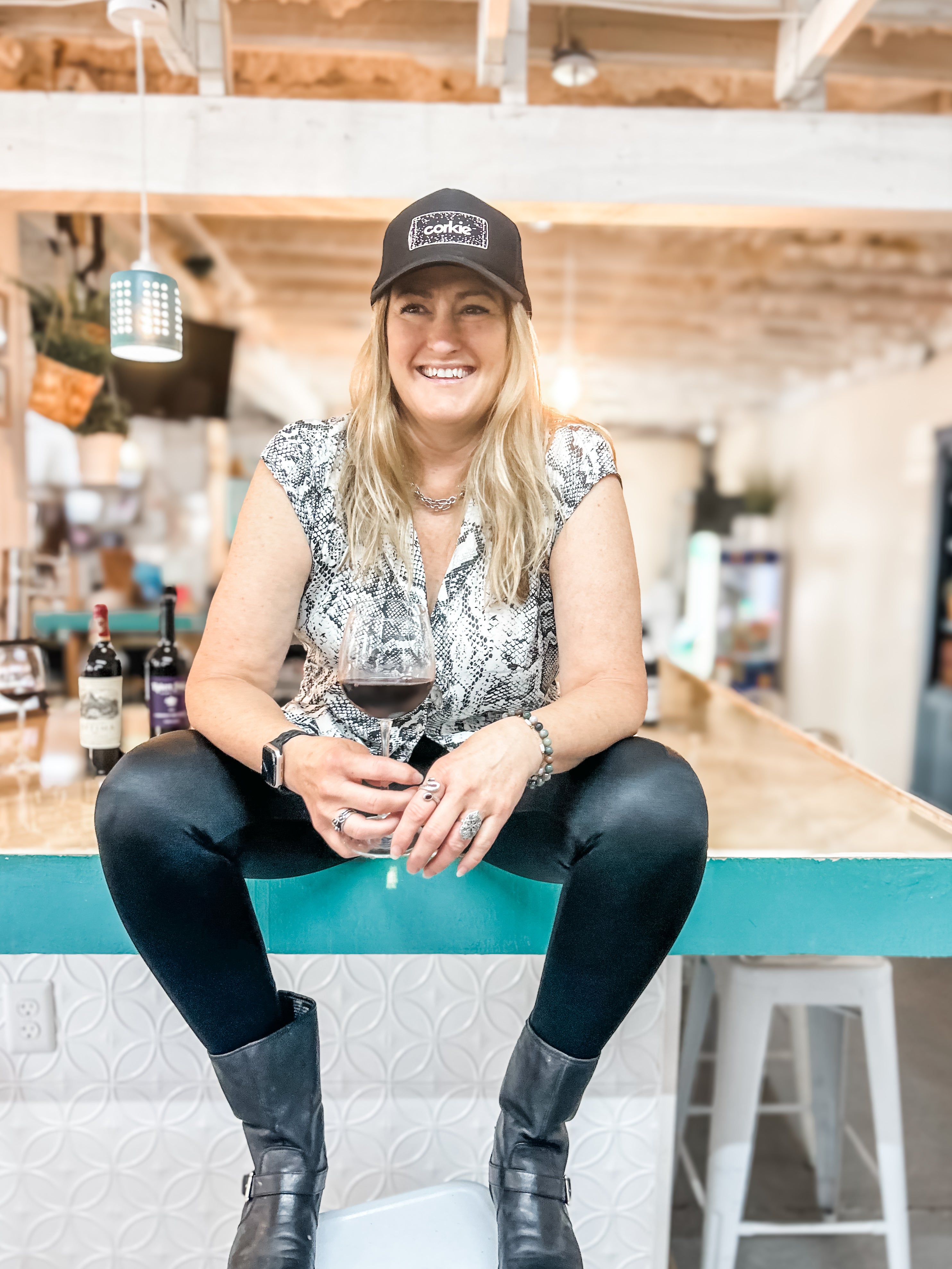 Woman sitting at a bar holding a glass of red wine, wearing a glitter corkie cork cap and black pants.