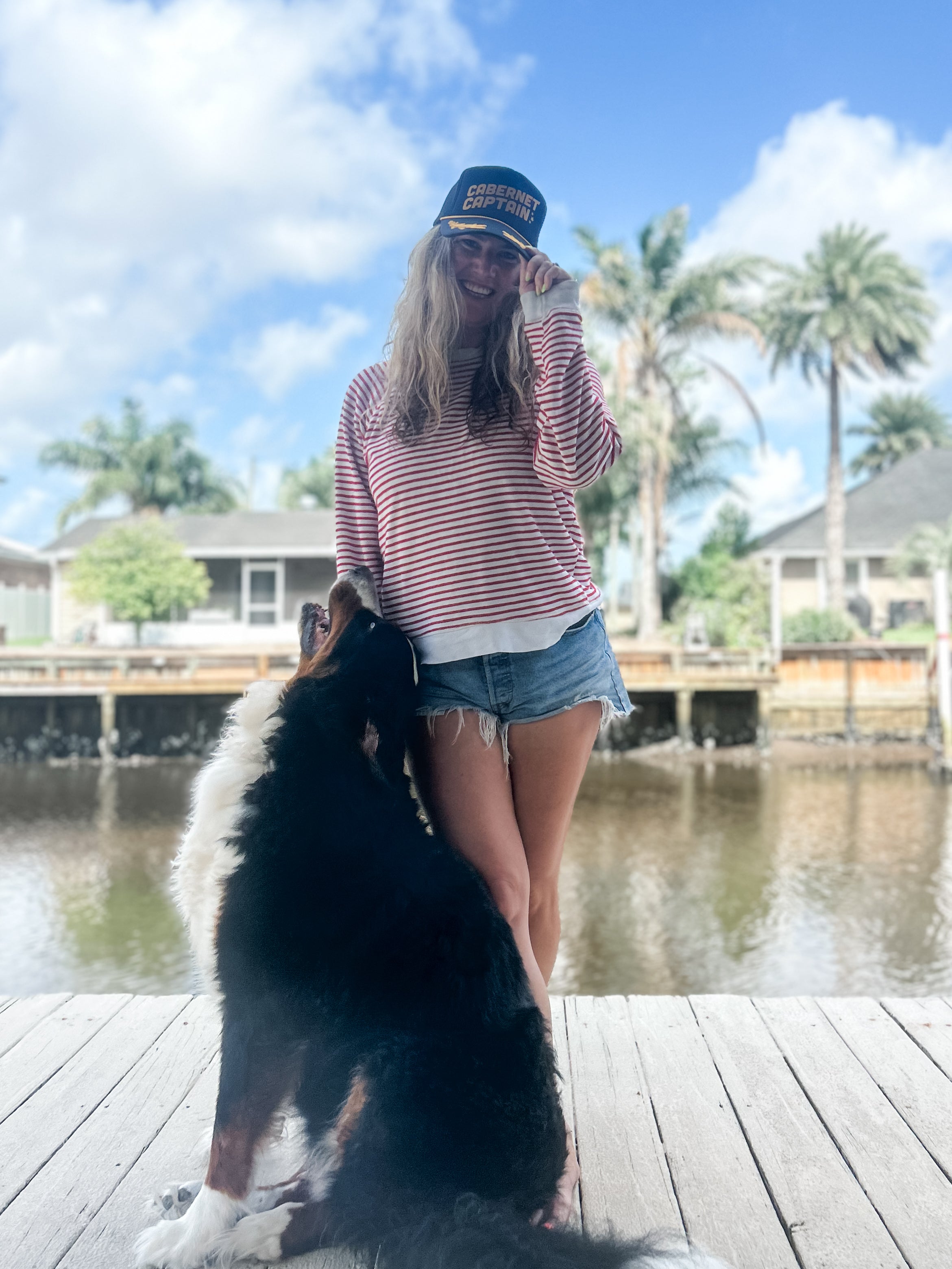 woman long hair standing on dock with nautical Cabernet captain corkie wine apparel black trucker hat and Clingy Bernese mountain dog