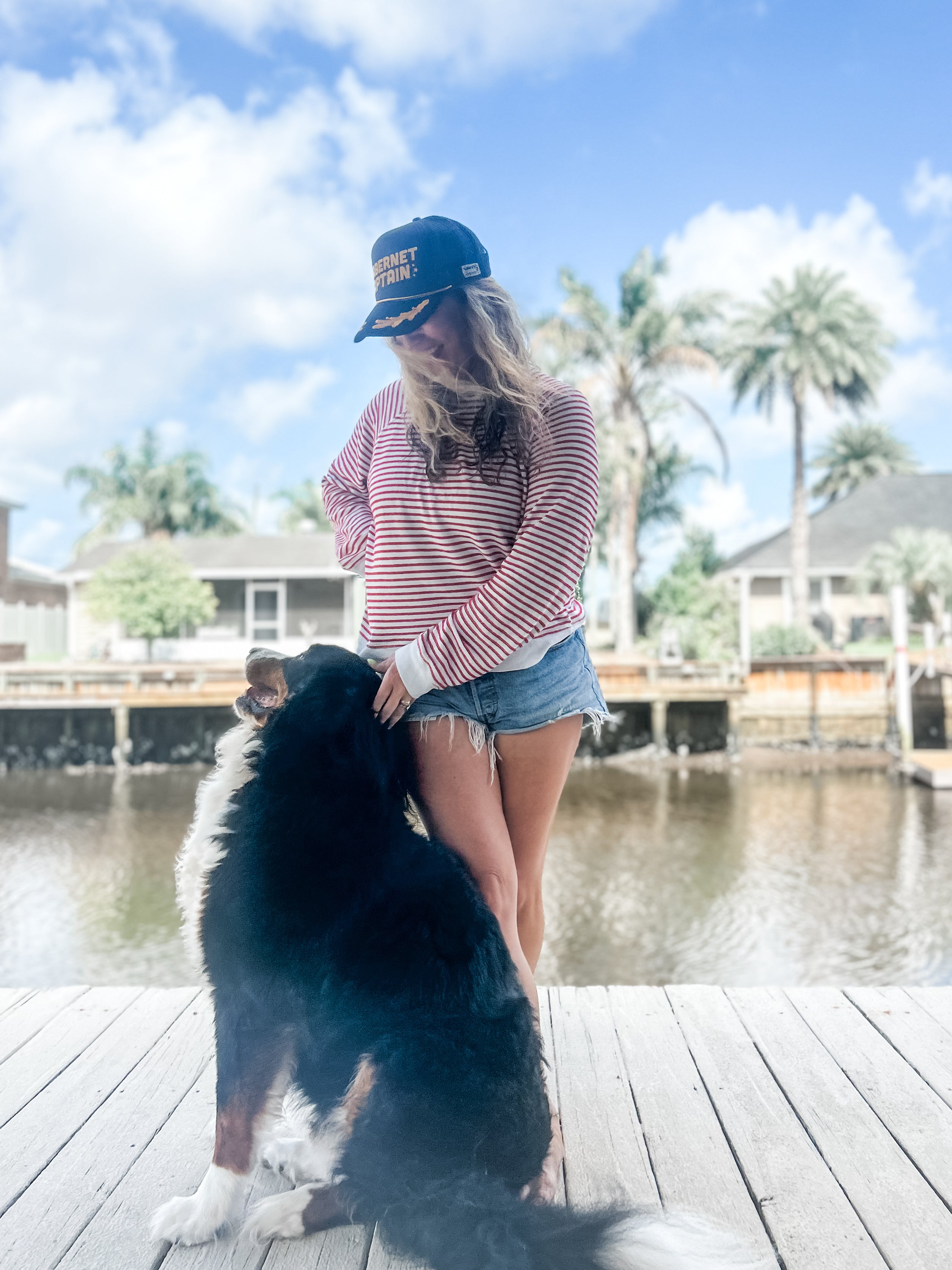 woman long hair standing on dock with nautical Cabernet captain corkie wine apparel black trucker hat and Bernese mountain dog