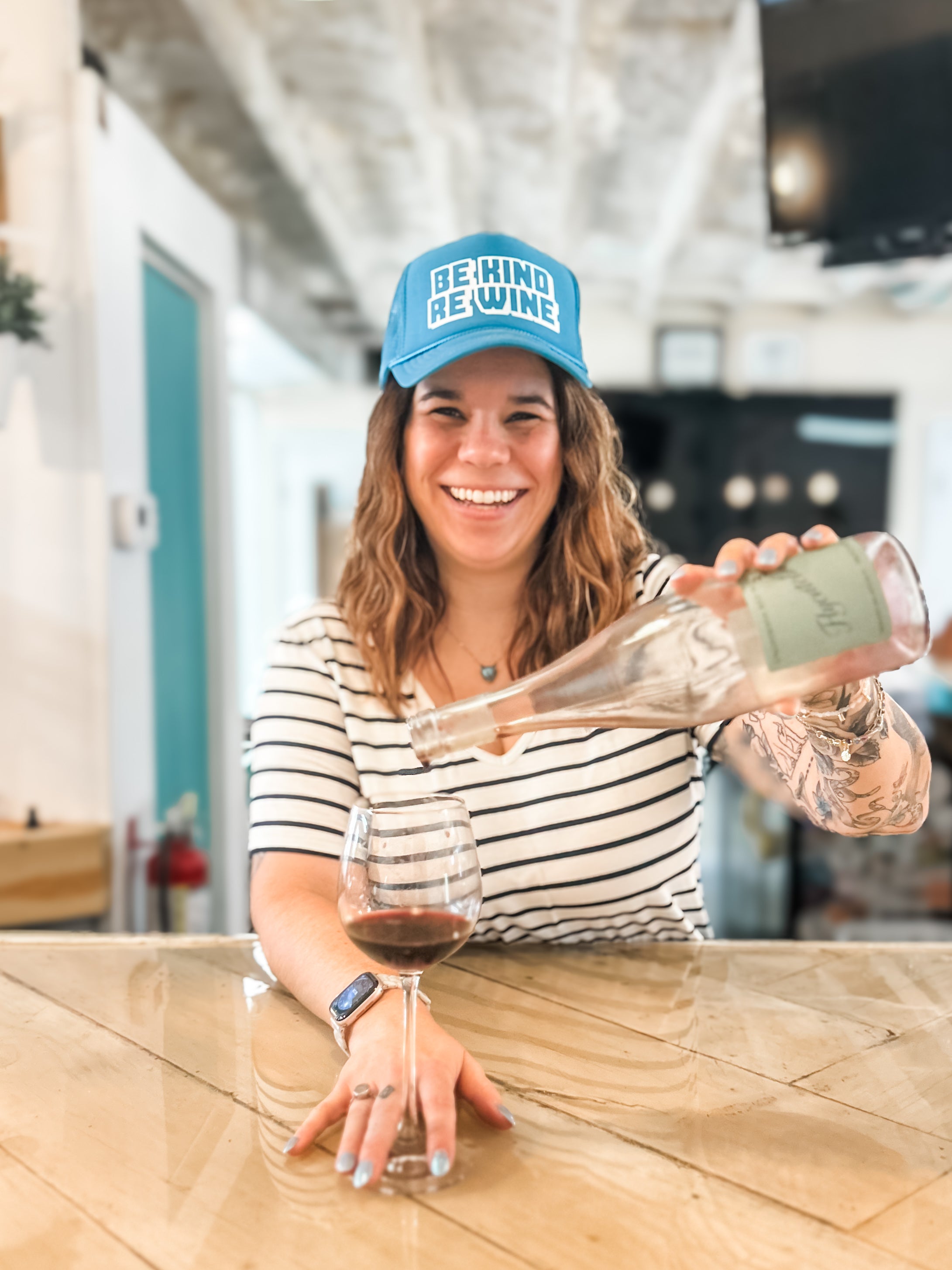 Woman pouring wine into a glass, wearing a 'be kind re Wine' corkie foam trucker cap.