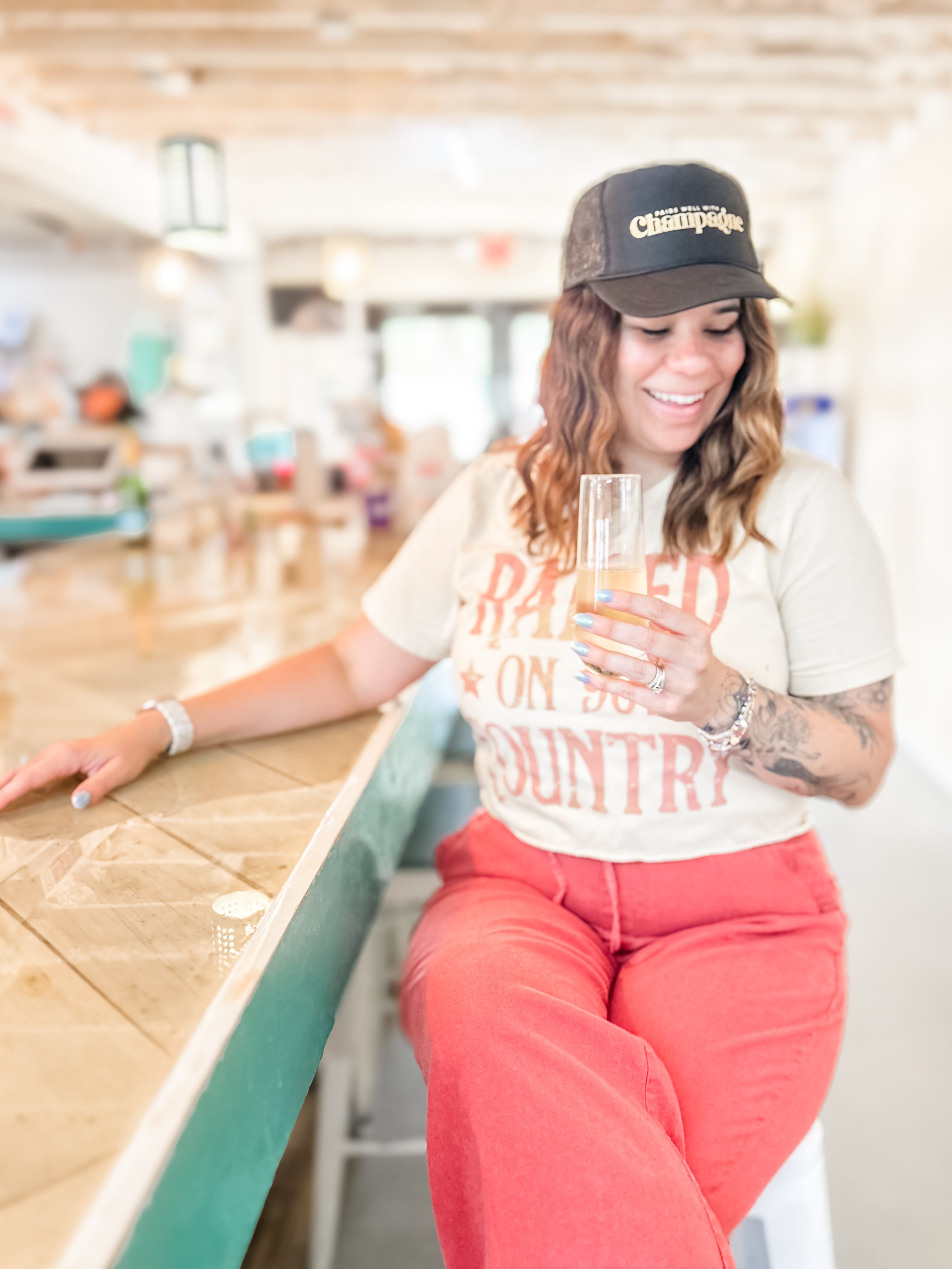 Woman sitting at a bar, wearing black pairs well with champagne foam corkie trucker hat, holding a glass of sparkling wine