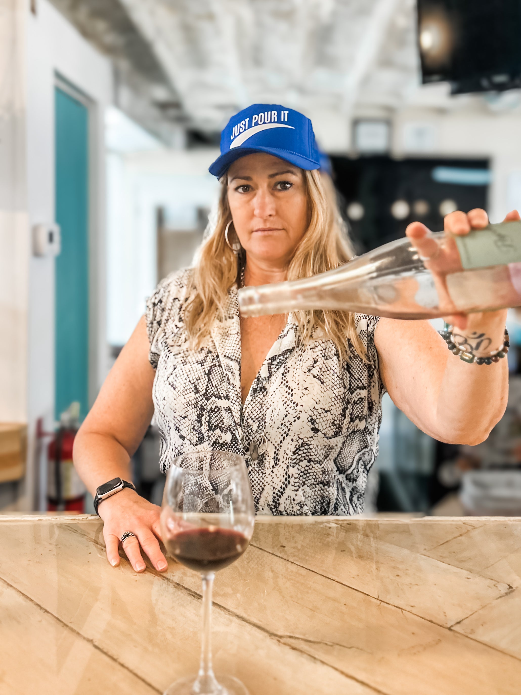 woamn pouring wine into a glass with a blue corkie foam cap labeled 'Just Pour It' in a bar setting.