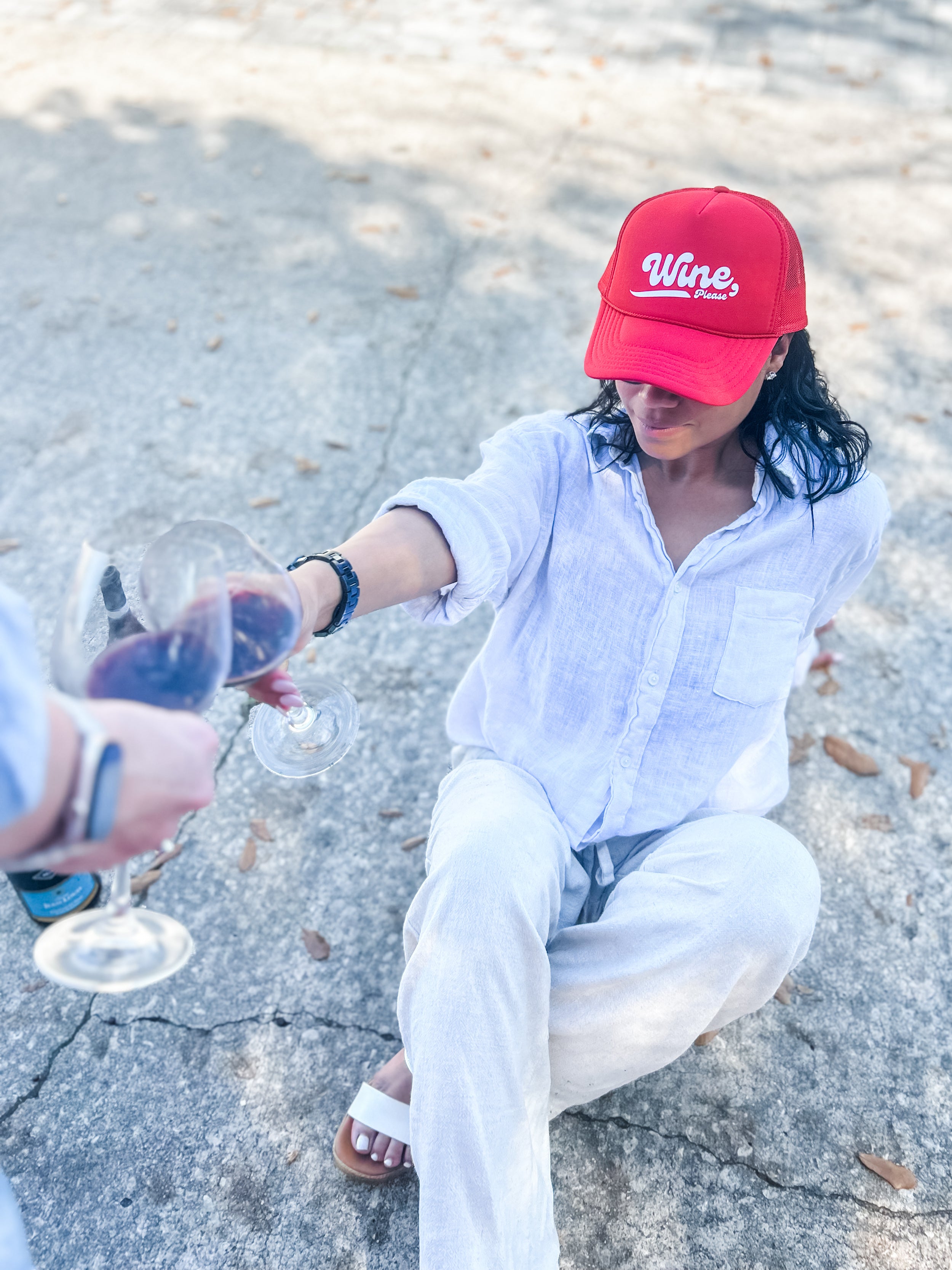woman wearing a red corkie 'Wine please' cap, pouring wine into a glass on a concrete surface.