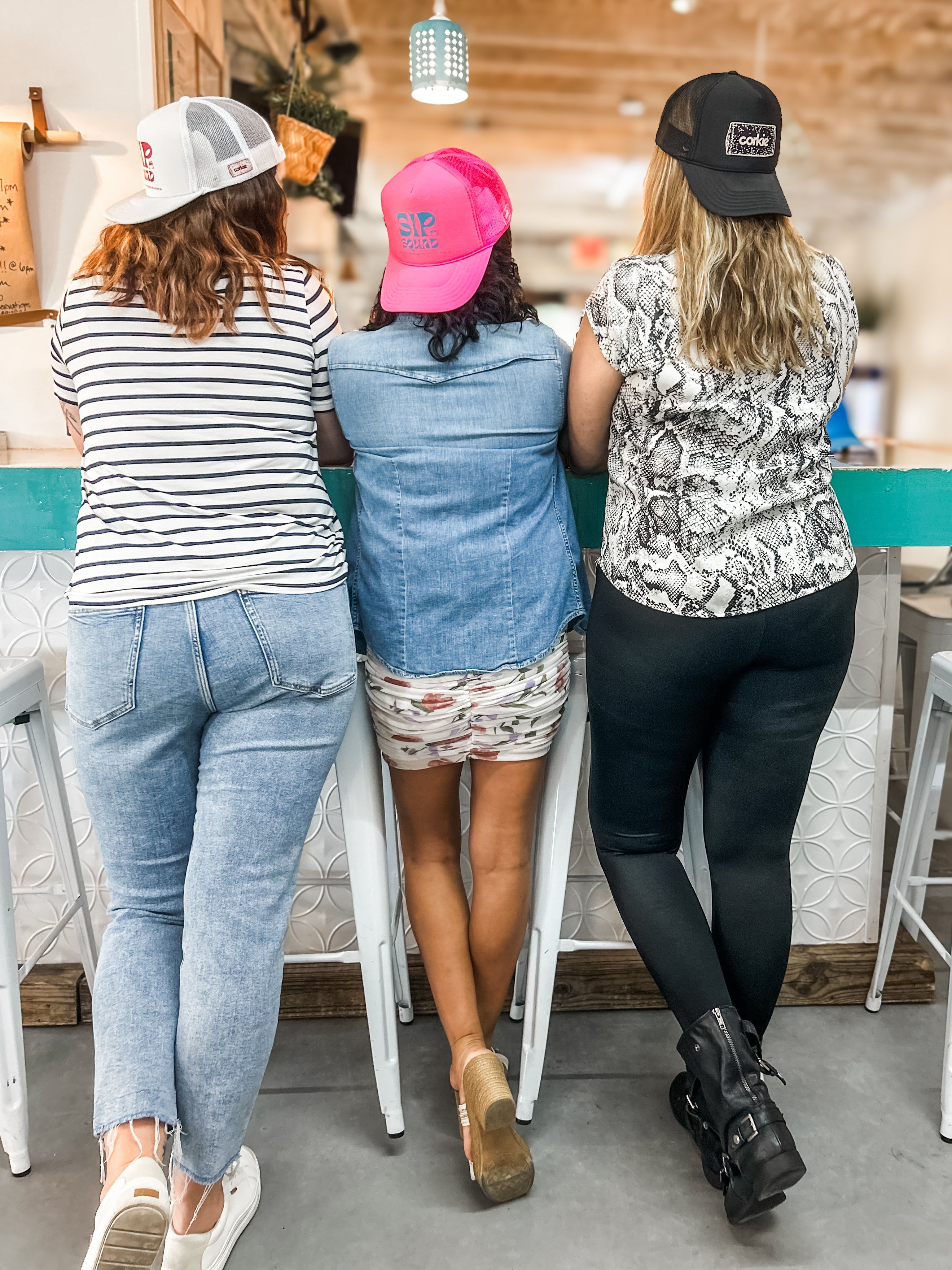 Three women standing together in a bar, wearing corkie hats saying Sip Squad, friends don't let friends sip alone