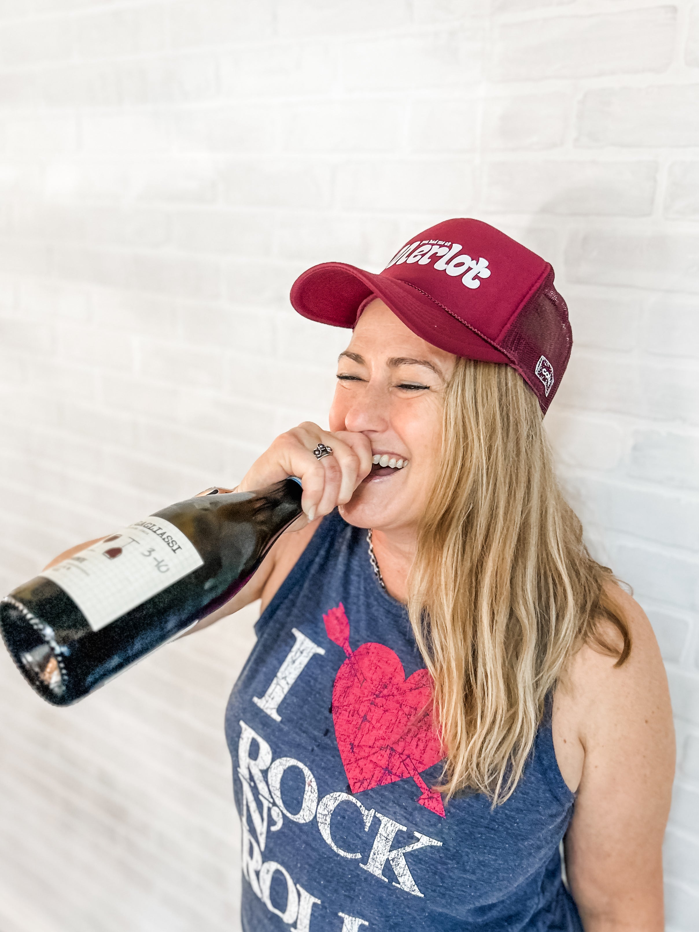 woman wearing a red corkie you had me at merlot cap and blue tank top with text, drinking a bottle.